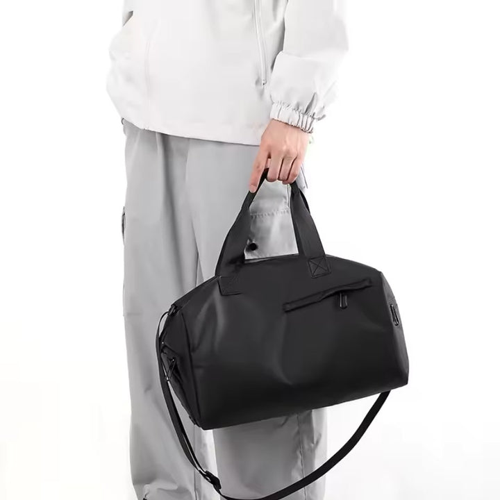 Petit sac de voyage homme noir porté à la main, bandoulière amovible et poche zippée pour accessoires.
