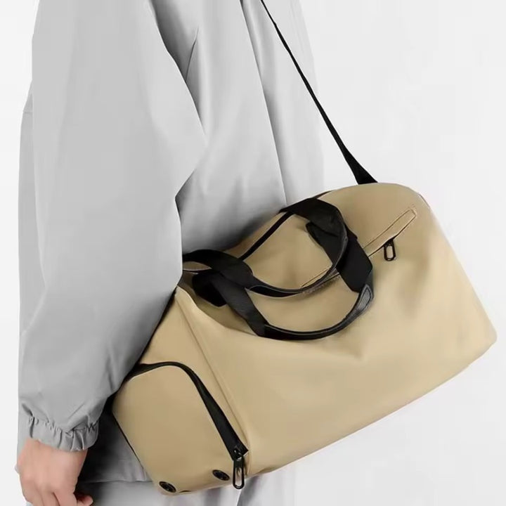 Petit sac de voyage homme beige porté à l’épaule, compartiment chaussures zippé compatible bagage cabine.