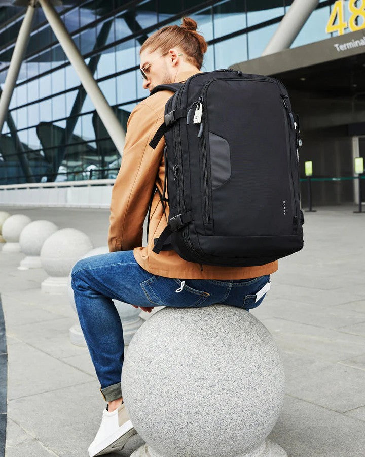Sac à Dos 55 x 35 x 25 cm - Format Bagage Cabine - porté sur le dos d'un homme assis sur une pierre devant le terminal d'un aéroport